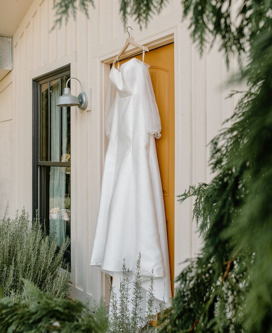 A white wedding dress hangs on a hanger outside a cottage door among greenery