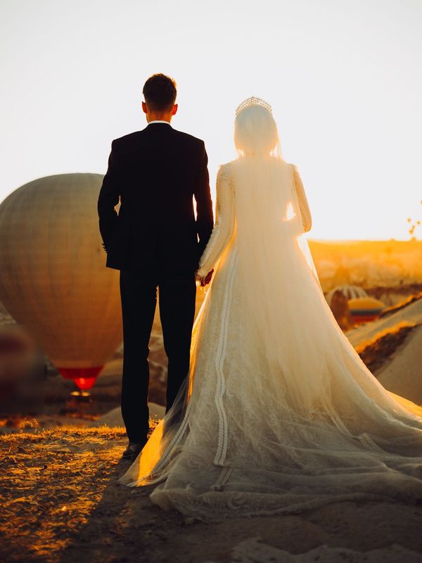 A bride in a long lace gown and veil and a groom in a dark suit hold hands at sunrise, seen from behind, with hot air balloons in the distance