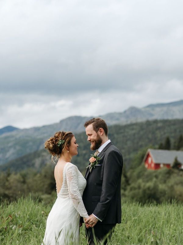 A bride and groom face each other and hold hands in a tall green meadow with mountains behind them