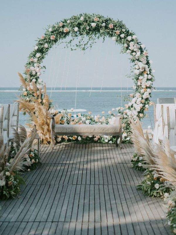 A round arch of white and blush flowers at the end of a boardwalk aisle lined with white chairs, with the ocean behind it